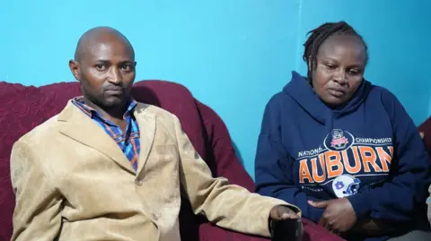Njoki's mother and father sit in front of a bright blue wall - her mother wears a blue hoody and father wears a beige blazer with a checked shirt. 